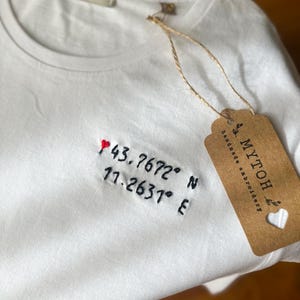 May include: White t-shirt with embroidered coordinates and a small red heart. The coordinates are 43.7672° N and 11.2631° E. A brown tag with the text "MYTOH handmade embroidery" is attached with twine.