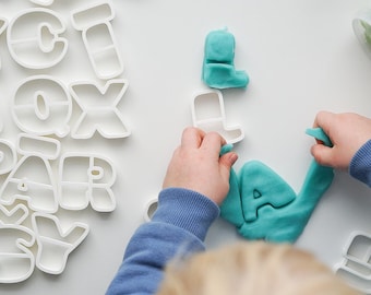 May include: A child uses cookie cutters shaped like letters to make play dough letters. The letters are 'L', 'A', and 'J'. The play dough is a light blue color.