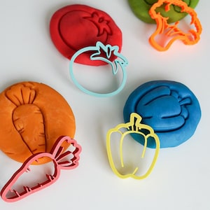May include: Five different colored play doughs with matching cookie cutters in the shapes of a tomato, carrot, bell pepper, broccoli, and a carrot. The cookie cutters are all different colors.