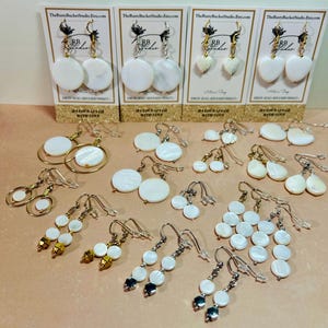 May include: A collection of handcrafted white shell earrings in various designs, including round, heart, and teardrop shapes. Some earrings feature gold-colored accents. The earrings are displayed on small cards with the text "TheRustyBucketStudio.Etsy.com".