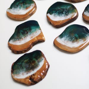 Puede incluir: Varios posavasos de madera con un diseño de resina que representa una escena de playa. Los posavasos presentan un océano verde azulado oscuro, olas blancas y una orilla arenosa de color canela. La madera tiene un borde natural y rústico.