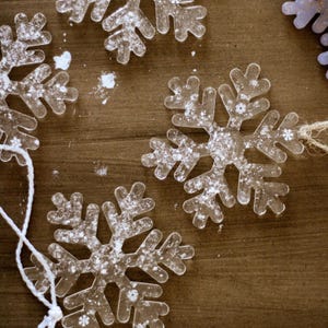 以下が含まれることがあります： 白いグリッターアクセントが付いた透明な雪の結晶オーナメントが木製の表面に散らばっています。白い紐または紐が付いている雪の結晶もあります。オーナメントは冬の休日のテーマです。