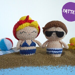 May include: Two crocheted figures, a girl with red and yellow hair wearing a blue bikini and a boy with brown hair wearing sunglasses and blue and white swim trunks, standing on a bed of gold glitter. The girl is holding a beach ball and the boy is holding a yellow rubber duck.