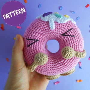May include: A pink crocheted donut with brown icing and colorful sprinkles. The donut has a happy face with black eyes and a smile.