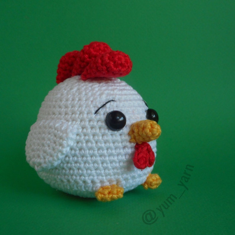 May include: White crocheted chicken toy with red comb, yellow beak, and black eyes. The toy is sitting on a green surface.