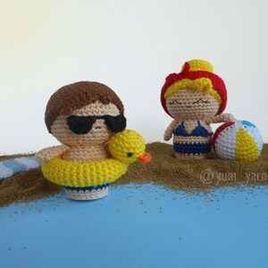 May include: Two crocheted figures on a beach scene. One figure is wearing sunglasses and a yellow life preserver. The other figure is wearing a blue swimsuit and holding a beach ball.