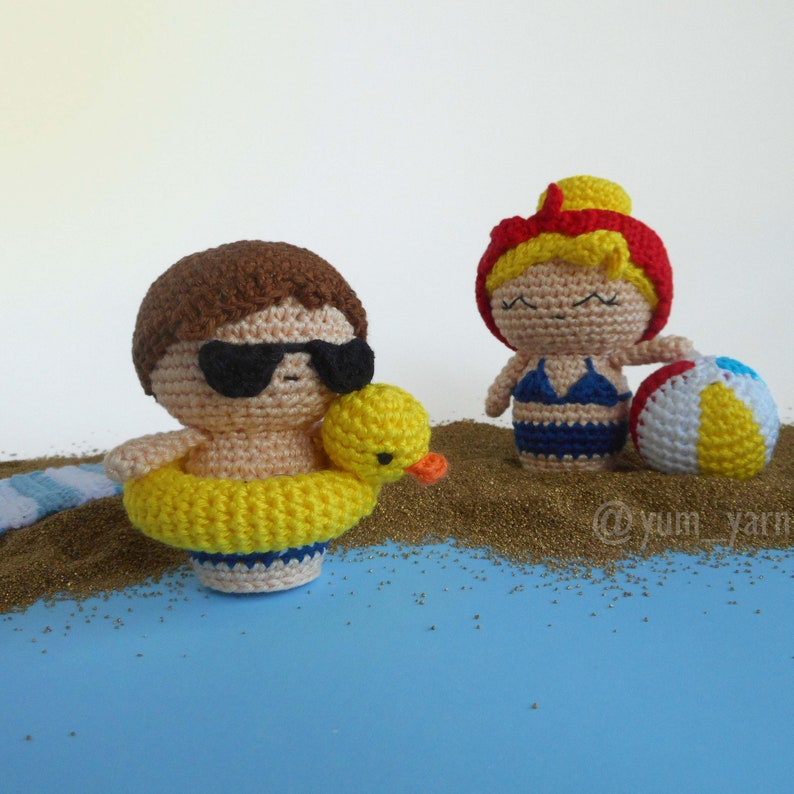 May include: Two crocheted figures on a beach scene. One figure is wearing sunglasses and a yellow life preserver. The other figure is wearing a blue swimsuit and holding a beach ball.