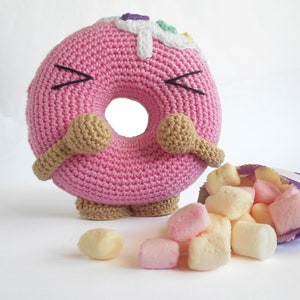 May include: A pink crocheted donut with brown arms and legs, with white frosting and colorful sprinkles. The donut is looking at a bag of pink and white marshmallows that have spilled out.
