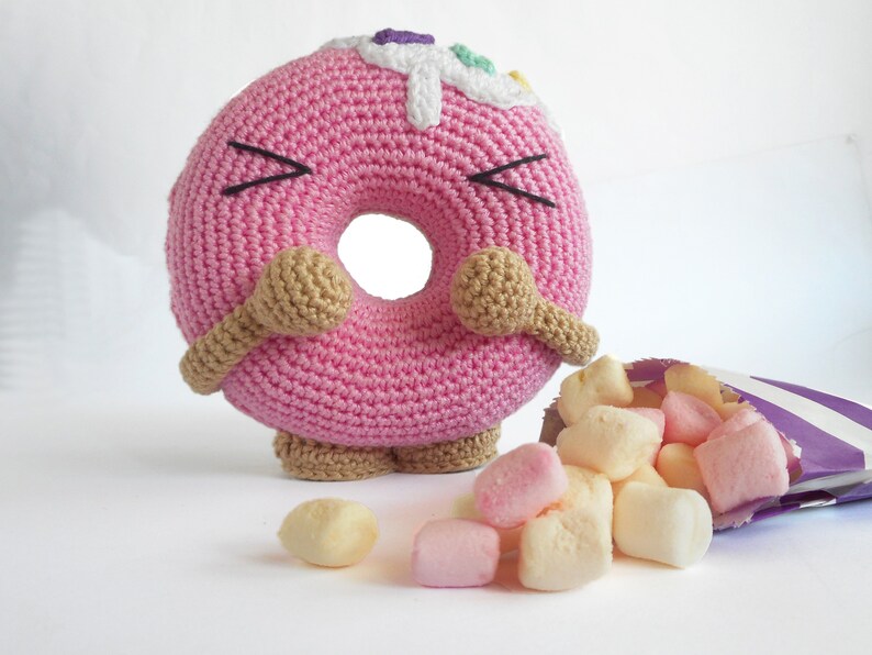 May include: A pink crocheted donut with brown arms and legs, with white frosting and colorful sprinkles. The donut is looking at a bag of pink and white marshmallows that have spilled out.