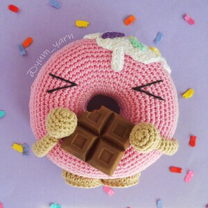 May include: A pink crocheted donut with a white frosting and colorful sprinkles, holding a chocolate bar in its arms. The donut has a cute face with black eyes and a smile.