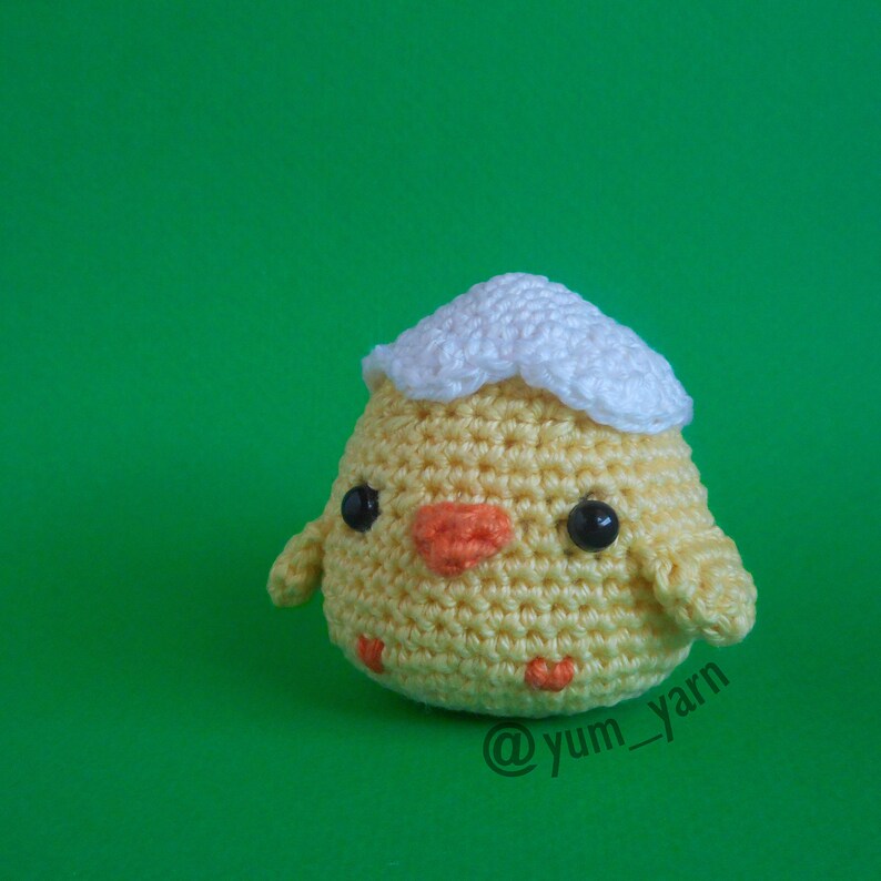 May include: A crocheted yellow chick with a white crocheted eggshell on its head. The chick has black eyes, an orange beak, and a small orange wattle. The chick is sitting on a green surface. The image is captioned with the text "@yum_yarn".