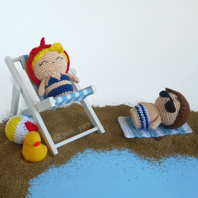 May include: Two crocheted dolls relaxing on a beach. One doll is wearing a blue swimsuit and laying on a white and blue striped beach chair. The other doll is wearing blue and white striped swim trunks and laying on a blue and white striped towel. Both dolls have brown eyes and are wearing sunglasses. The dolls are surrounded by brown sand and a blue surface.