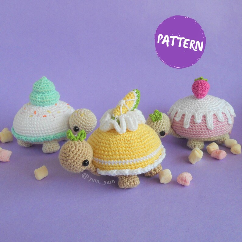 Op de afbeelding: Drie gehaakte schildpad speelgoedjes die zijn versierd om eruit te zien als cupcakes. De schildpadden zijn gemaakt van beige, wit, roze, geel en groen garen. De cupcakes zijn bedekt met een groene swirl, een citroenschijfje en een roze aardbei. De schildpadden zitten op een paarse ondergrond met witte marshmallows die eromheen zijn verspreid. De afbeelding bevat de tekst "PATTERN".