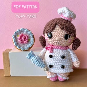 Puede incluir: Muñeca de chef de ganchillo con cabello castaño, gorro blanco y bufanda rosa, sosteniendo una manga pastelera azul. También hay un pastelito de ganchillo. El texto "PDF PATTERN" y "Yum Yarn" son visibles.
