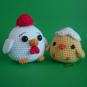 May include: A white crocheted chicken with a red comb and a yellow beak, next to a yellow crocheted chick in a white eggshell.