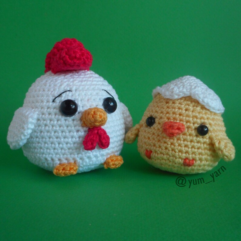 May include: A white crocheted chicken with a red comb and a yellow beak, next to a yellow crocheted chick in a white eggshell.