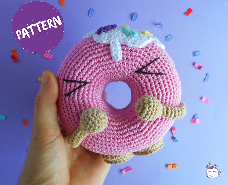May include: A pink crocheted donut with brown icing and colorful sprinkles. The donut has a happy face with black eyes and a smile.