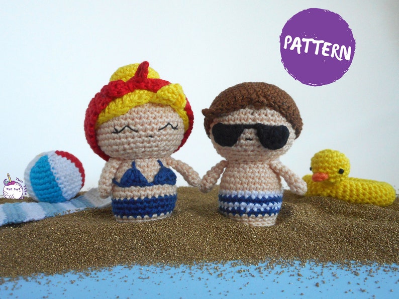 May include: Two crocheted figures, a girl with red and yellow hair wearing a blue bikini and a boy with brown hair wearing sunglasses and blue and white swim trunks, standing on a bed of gold glitter. The girl is holding a beach ball and the boy is holding a yellow rubber duck.
