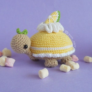 Op de afbeelding: Een gehaakte schildpad die een geel en wit gehaakt schild draagt met een citroenschijf erop. De schildpad is omringd door witte en roze marshmallows. De afbeelding bevat de tekst "@yum_yarn".