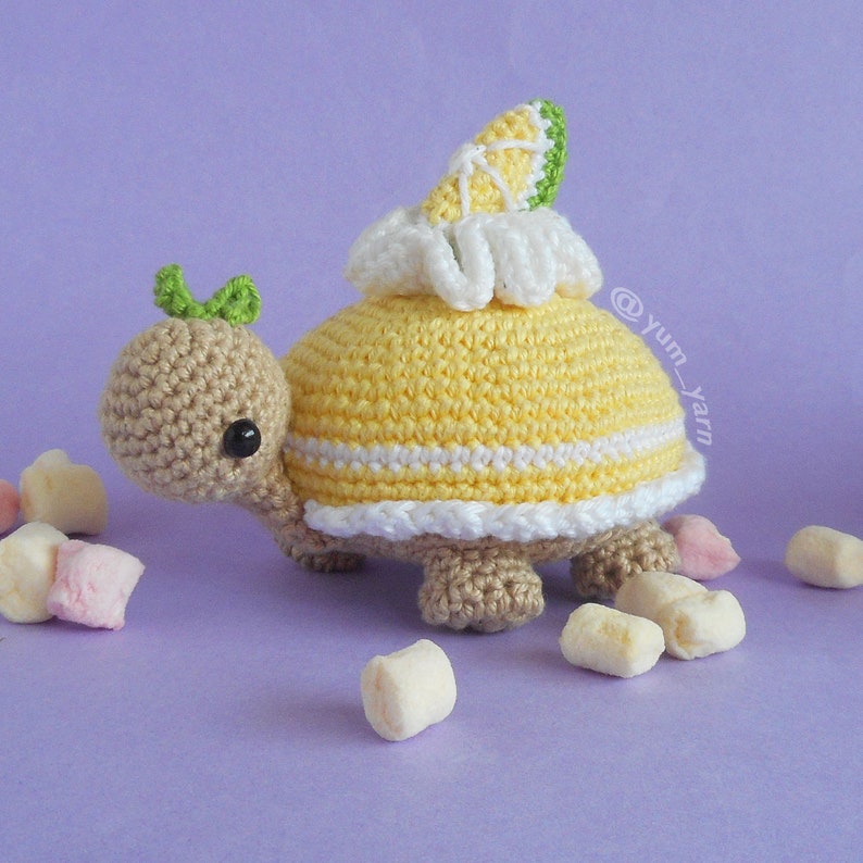 Op de afbeelding: Een gehaakte schildpad die een geel en wit gehaakt schild draagt met een citroenschijf erop. De schildpad is omringd door witte en roze marshmallows. De afbeelding bevat de tekst "@yum_yarn".