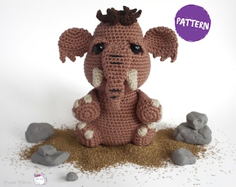 Könnte beinhalten: Ein gehäkeltes braunes Mammut-Spielzeug, das auf einem Bett aus goldenem Glitzer mit grauen Steinen drumherum sitzt. Das Spielzeug hat eine braune und schwarze Mähne und große schwarze Augen. Der Text "PATTERN" befindet sich auf einem lila Kreis in der oberen rechten Ecke des Bildes.