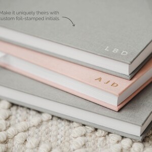 May include: A stack of three hardcover journals with fabric covers in shades of gray and light pink. The top gray journal features the foil-stamped initials LBD, the middle pink journal has AJD, and the text Make it uniquely theirs with custom foil-stamped initials is visible.