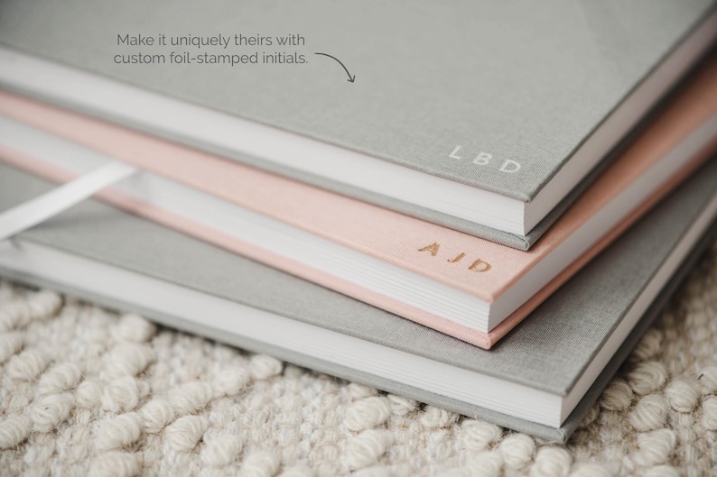 May include: A stack of three hardcover journals with fabric covers in shades of gray and light pink. The top gray journal features the foil-stamped initials LBD, the middle pink journal has AJD, and the text Make it uniquely theirs with custom foil-stamped initials is visible.