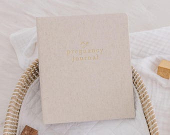 May include: A beige pregnancy journal with gold lettering, resting in a woven basket. The journal's cover features the words "by pregnancy journal". A wooden block sits nearby on a white cloth.