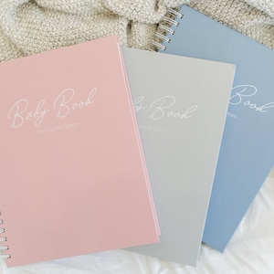 May include: Three spiral-bound journals in pink, gray, and blue. Each cover features the white script text Baby Book and the subtitle The first five years. The notebooks are arranged in a staggered stack, showcasing their matching metallic bindings.