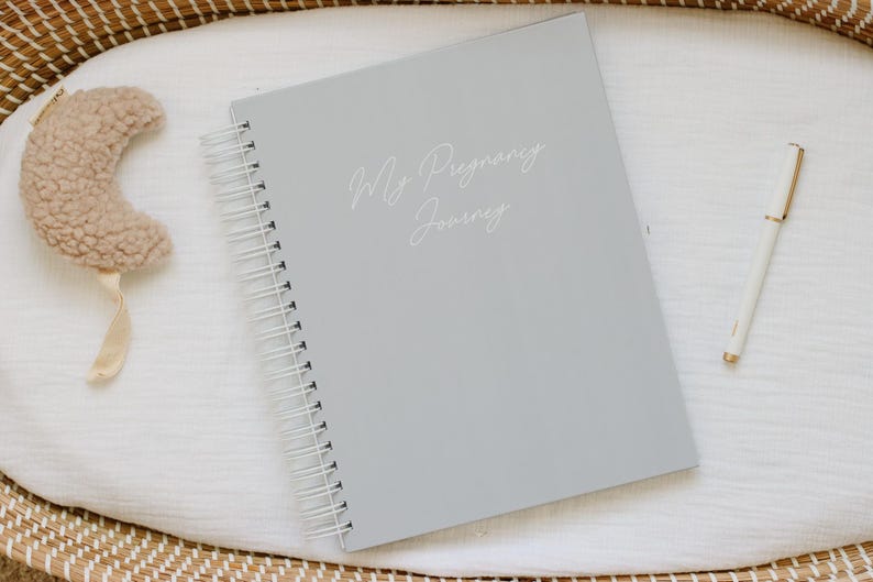 May include: A light gray spiral-bound journal with the text My Pregnancy Journey printed in white script on the cover. The journal is accompanied by a white pen and a small, textured, beige crescent moon-shaped object with a ribbon.