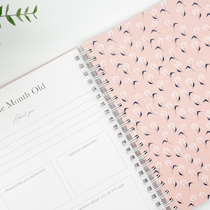 May include: Open baby memory book with spiral binding. The left page reads "One Month Old" with space to write. The right page has a pink floral pattern. The book includes sections for milestones and special memories.