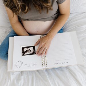 May include: A spiral-bound pregnancy journal with white pages featuring printed text including First Ultrasound and We're Having a Baby. A black and white ultrasound scan is placed on the left page, which has a floral wreath graphic.
