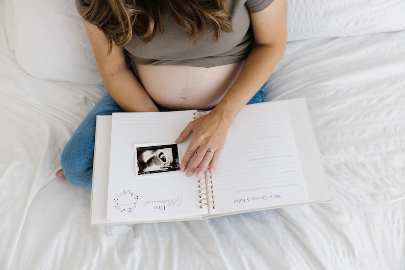 May include: A spiral-bound pregnancy journal with white pages featuring printed text including First Ultrasound and We're Having a Baby. A black and white ultrasound scan is placed on the left page, which has a floral wreath graphic.