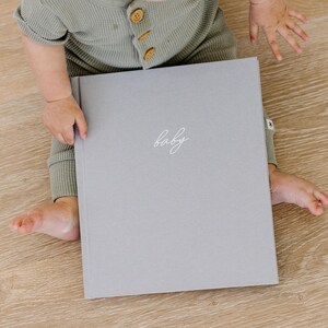 May include: A light gray hardcover book with the word baby written in white cursive script on the front cover. The book is held by a child wearing a sage green waffle knit long-sleeved romper with small brown buttons down the front chest area.
