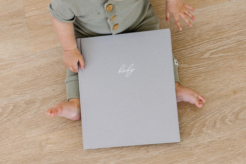 May include: A light gray hardcover book with the word baby written in white cursive script on the front cover. The book is held by a child wearing a sage green waffle knit long-sleeved romper with small brown buttons down the front chest area.