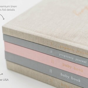 May include: A stack of four hardcover journals wrapped in linen fabric. The top journal is beige, followed by a grey journal labeled pregnancy journal, a pink baby book, and a grey baby book. Each spine features gold foil text and vertical lines.