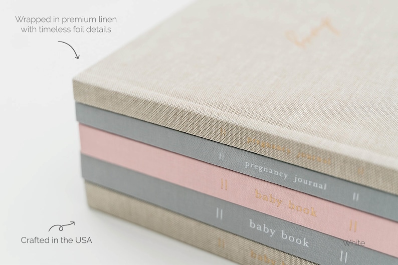 May include: A stack of four hardcover journals wrapped in linen fabric. The top journal is beige, followed by a grey journal labeled pregnancy journal, a pink baby book, and a grey baby book. Each spine features gold foil text and vertical lines.