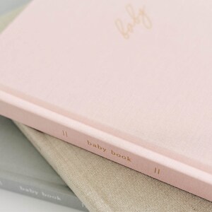 May include: A light pink hardcover book with the word baby written in gold cursive on the front cover and the text baby book printed in gold on the spine. The book is stacked on top of a tan book and a gray book, both featuring similar text.