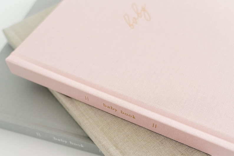 May include: A light pink hardcover book with the word baby written in gold cursive on the front cover and the text baby book printed in gold on the spine. The book is stacked on top of a tan book and a gray book, both featuring similar text.
