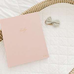May include: A light pink hardcover book with the word baby written in gold cursive script on the front cover and the initials B.M.F. in gold at the bottom right corner, accompanied by a small sage green fabric bow accessory.