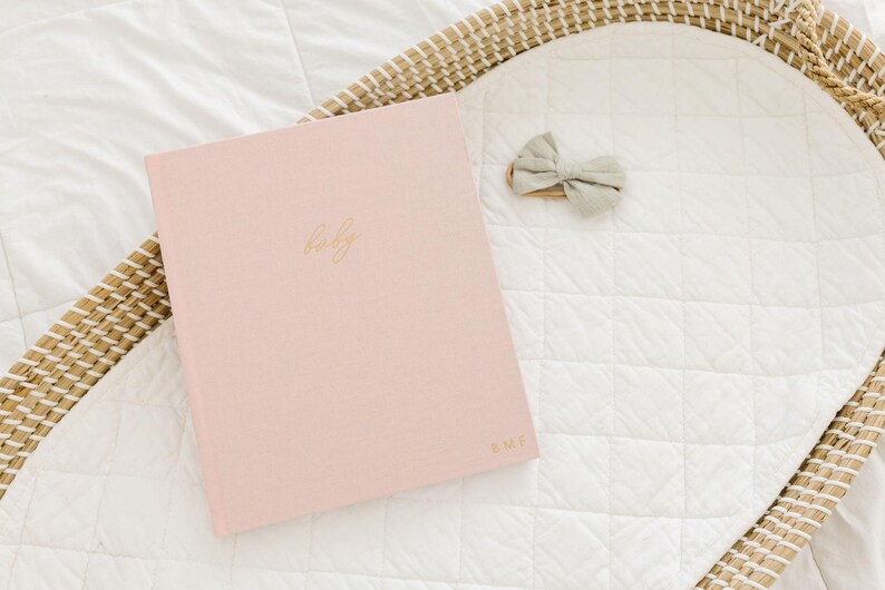 May include: A light pink hardcover book with the word baby written in gold cursive script on the front cover and the initials B.M.F. in gold at the bottom right corner, accompanied by a small sage green fabric bow accessory.