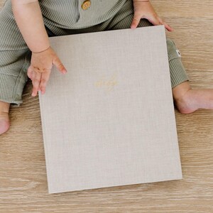 May include: A rectangular photo album with a light beige linen cover and gold embossed cursive text centered on the front. The album is held by a person wearing sage green waffle-knit clothing, showcasing the textured fabric and neutral color.