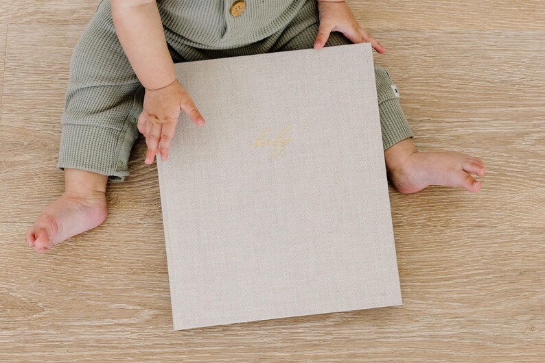 May include: A rectangular photo album with a light beige linen cover and gold embossed cursive text centered on the front. The album is held by a person wearing sage green waffle-knit clothing, showcasing the textured fabric and neutral color.
