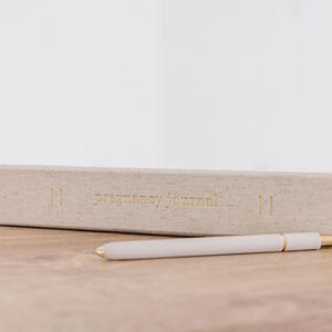 May include: A light beige fabric-covered pregnancy journal with gold foil lettering on the spine that reads pregnancy journal, accompanied by a matching light beige pen with gold-colored metal accents positioned horizontally in front of the book.