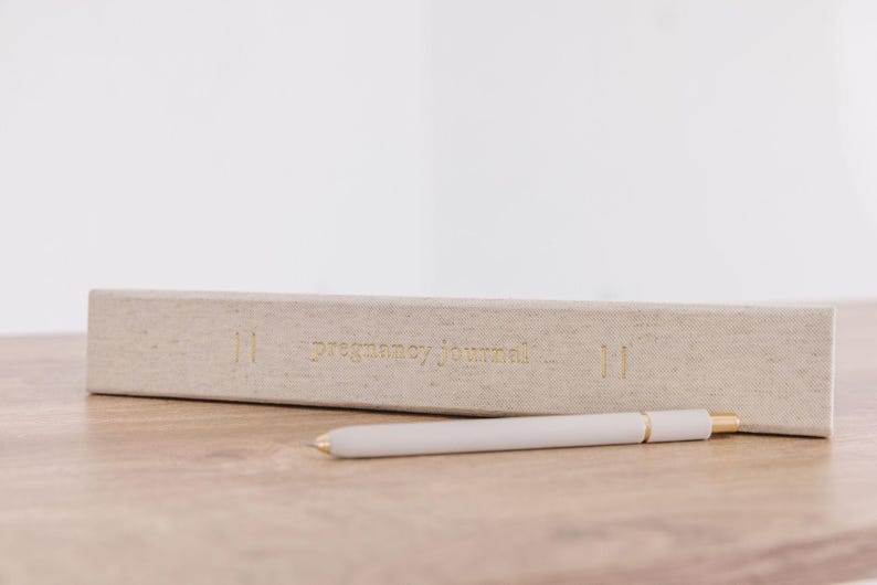 May include: A light beige fabric-covered pregnancy journal with gold foil lettering on the spine that reads pregnancy journal, accompanied by a matching light beige pen with gold-colored metal accents positioned horizontally in front of the book.