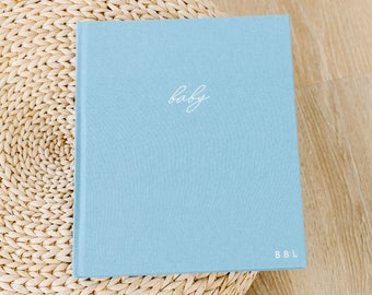 May include: A light blue baby memory book with the word "baby" in cursive script. The book has a linen texture and the initials "BBL" are at the bottom right corner. The book is on a woven, circular surface.