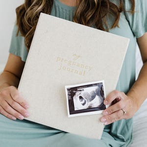 May include: A light beige, fabric-covered pregnancy journal with the text "my pregnancy journal" embossed in gold on the front cover. A small, rectangular black and white ultrasound scan is positioned on the lower center of the journal cover.