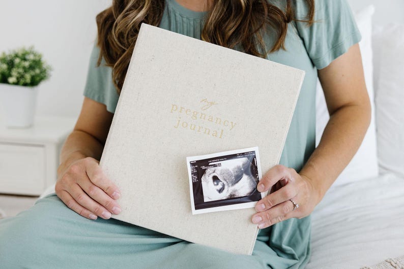 May include: A light beige, fabric-covered pregnancy journal with the text "my pregnancy journal" embossed in gold on the front cover. A small, rectangular black and white ultrasound scan is positioned on the lower center of the journal cover.