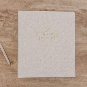 May include: A light beige, textured hardcover journal with the words "my pregnancy journal" embossed in gold lettering on the front cover, accompanied by a matching light beige pen with gold accents positioned to the left of the book.