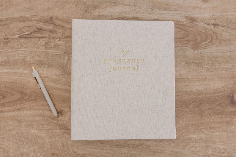 May include: A light beige, textured hardcover journal with the words "my pregnancy journal" embossed in gold lettering on the front cover, accompanied by a matching light beige pen with gold accents positioned to the left of the book.